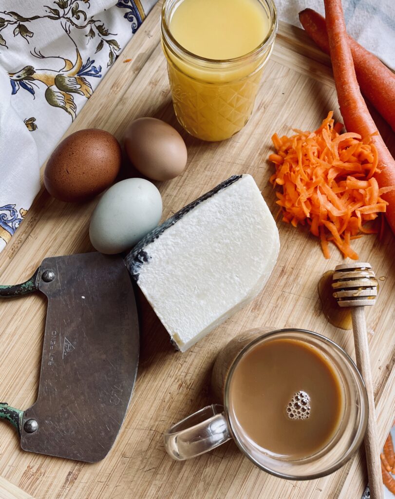 Cutting board with Raw Honey, Coffee with Cream, Shredded Carrots, Orange Juice, Eggs, knife and Raw Cheese on it.