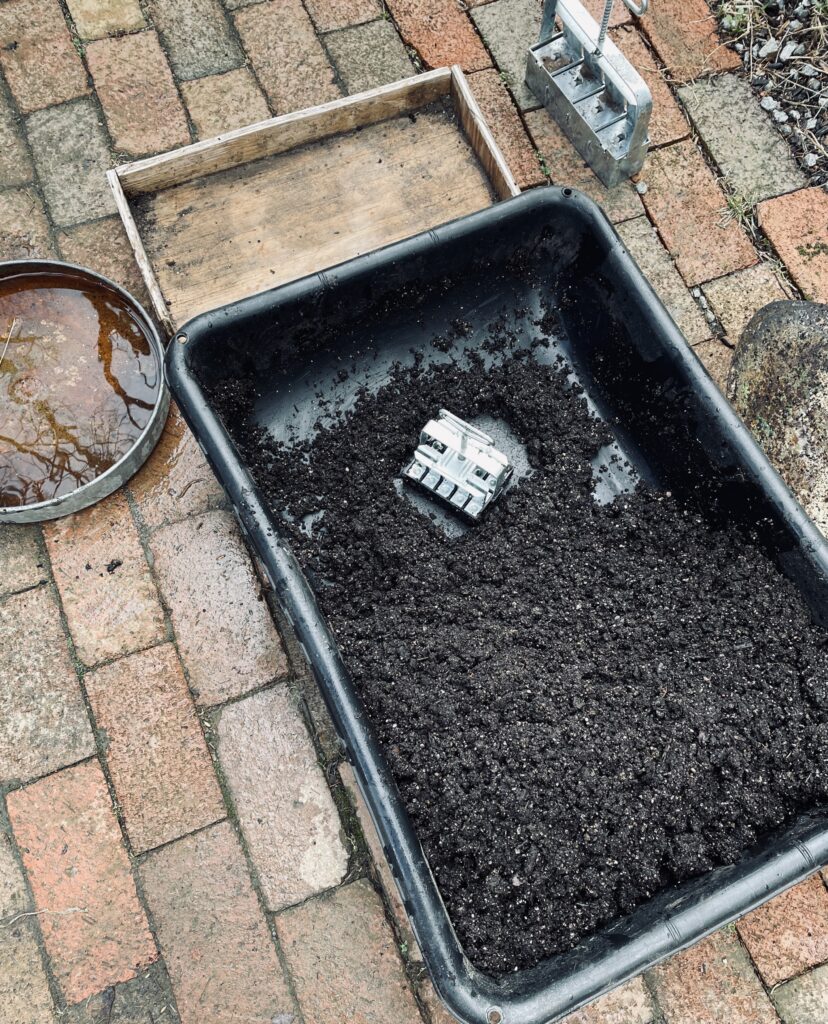 Black plastic tub full of soil, soil blocker, brick path, wooden tray