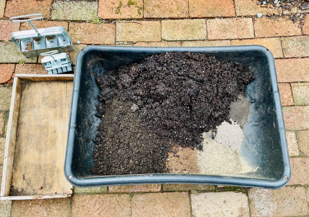 Black plastic tub with soil and amendments. Brick path. wooden tray and soil blockers