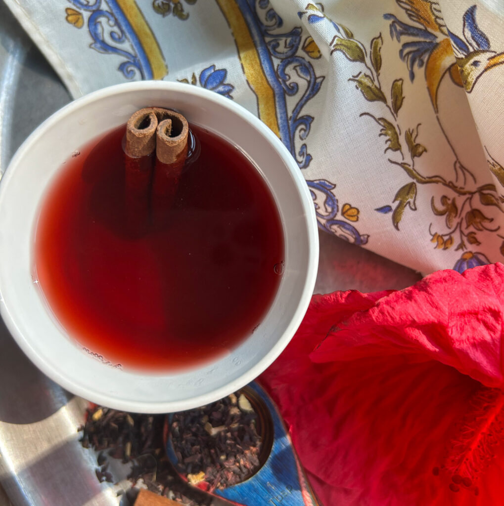 Overhead shot of a cup of tea in a white mug. Placed on a silver tray with a scoop of loose tea next to a fresh hibiscus flower and surrounded by a cloth napkin.