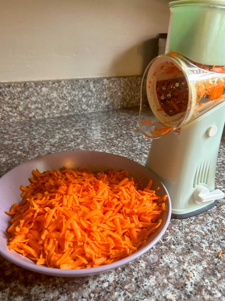 Vegetable grater grating raw carrots. Shredded carrots in purple bowl.