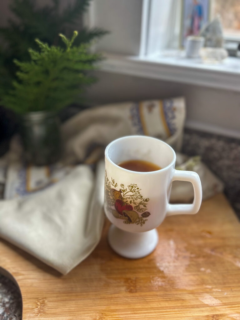 White mug with broth on top of cutting board. Printed napkin and evergreens in the background. 