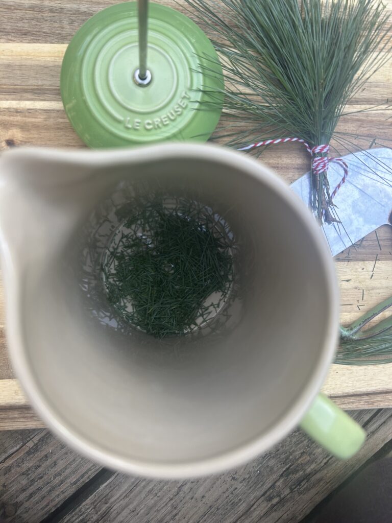 A green french press with the lid off and chopped white pine needles inside, on a cutting board with a bundle of white pine needles and an herb chopper.