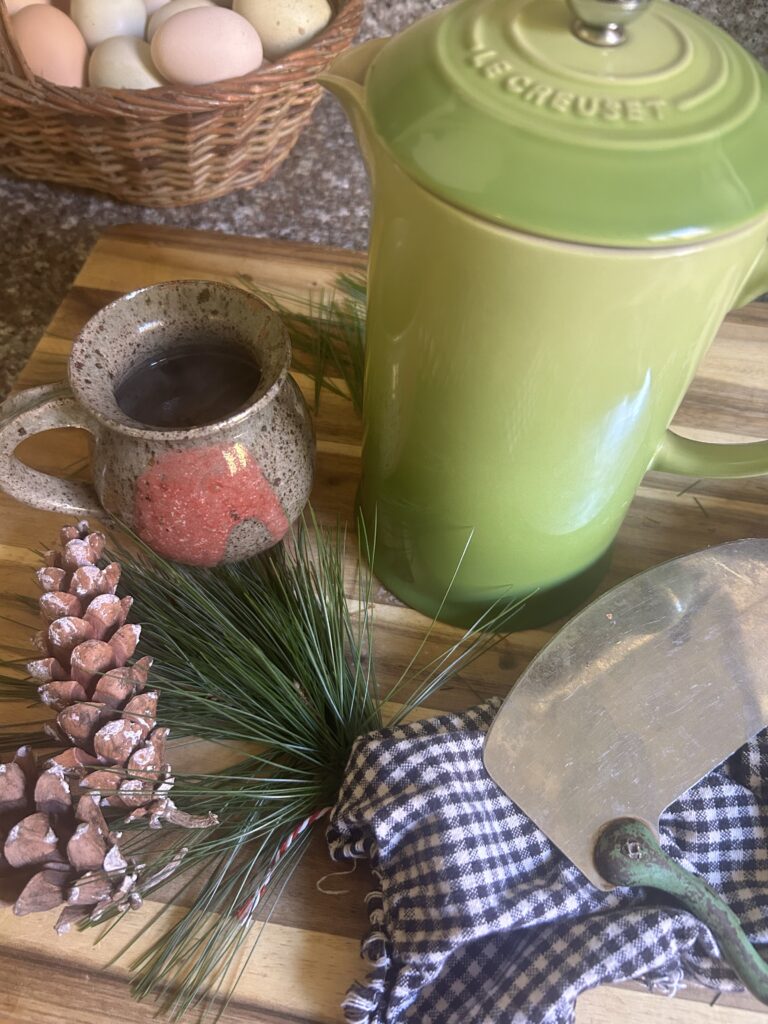 Cutting board with green french press, a brown and red tea mug, a bundle od white pine needles, 2 white pine cones and a black and white checkered napkin. With a basket of eggs in the background.